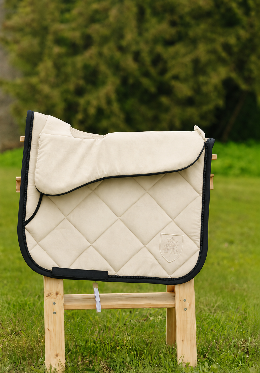 Ivory quilted dressage saddle pad with black cord piping and embroidered crest on a wooden stand